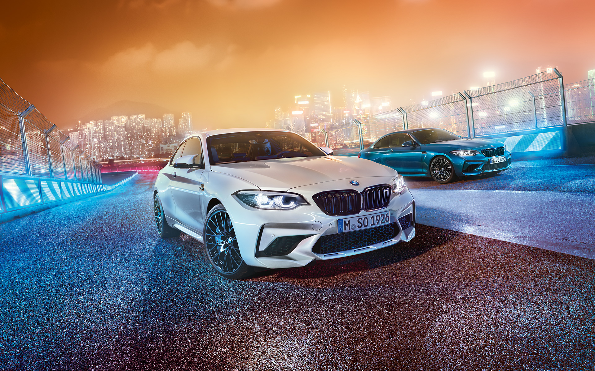 Two BMW M2 Competition on race track White and blue BMW M2 Competition standing on race track in front of nocturnal cityscape.