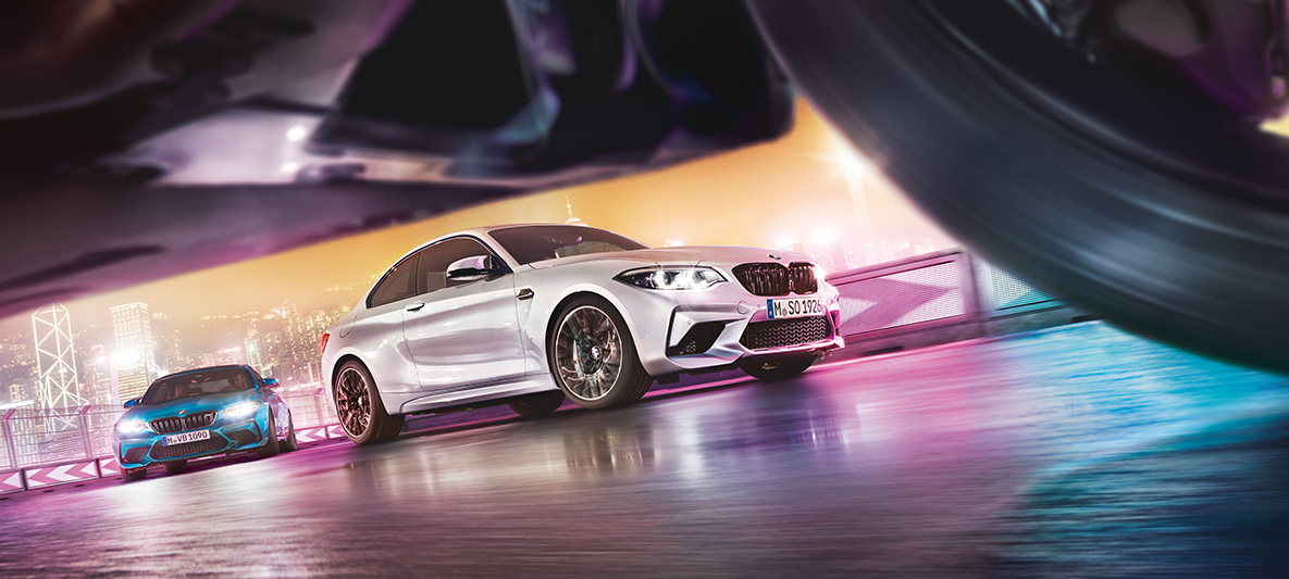 BMW M2 Competition on reflective asphalt. Two BMW M2 Competition on race track in front of nocturnal cityscape in street-level view.