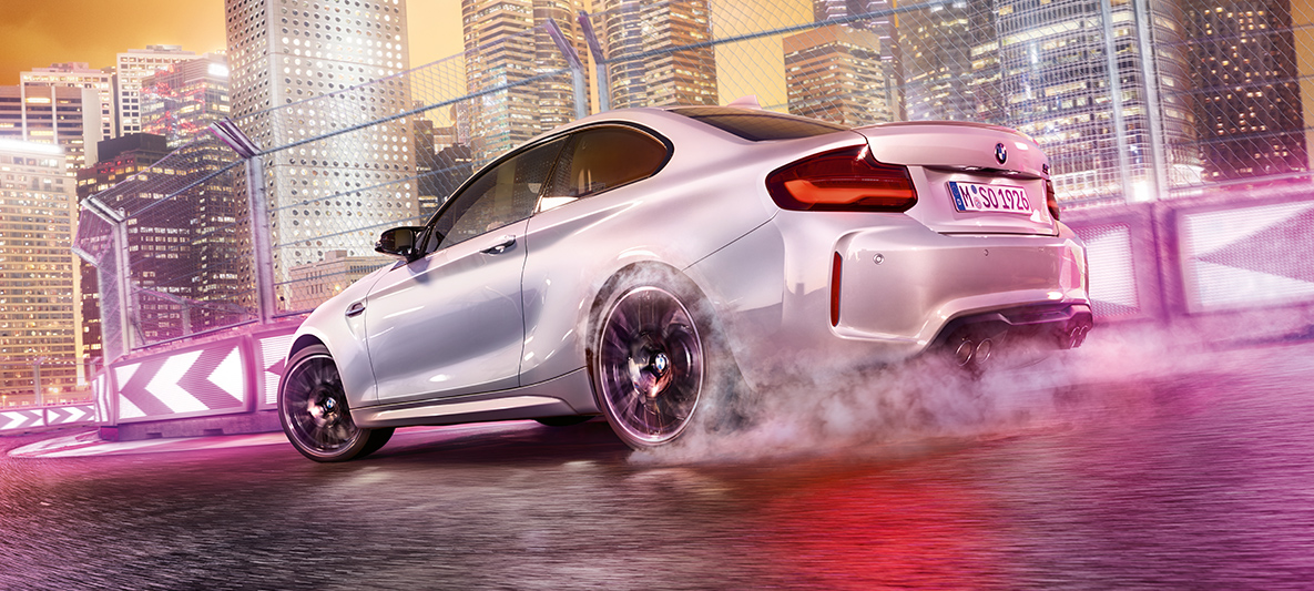 BMW M2 Competition accelerates White BMW M2 Competition drifting on race track in front of nocturnal skyline.